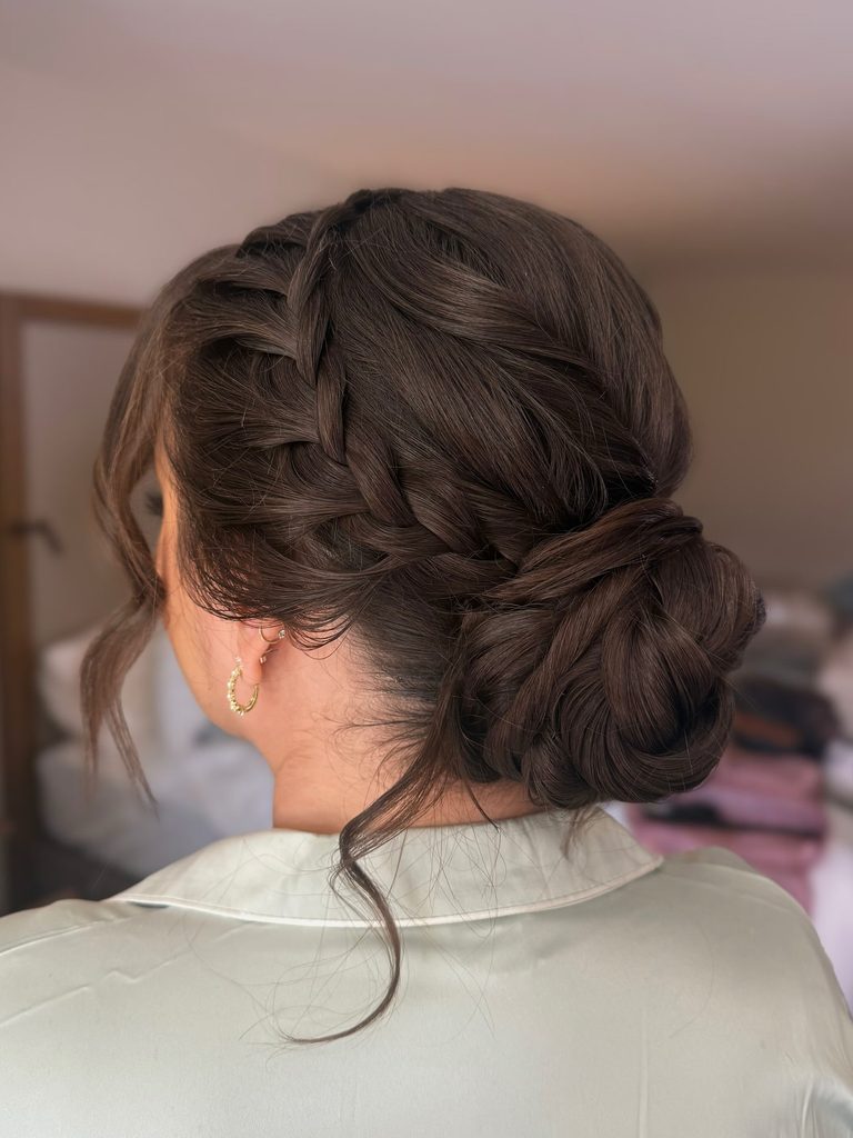 Side view of a woman with dark brunette hair styled in a French braid that leads into a twisted low bun with a few loose wispy strands near her ear and neck.