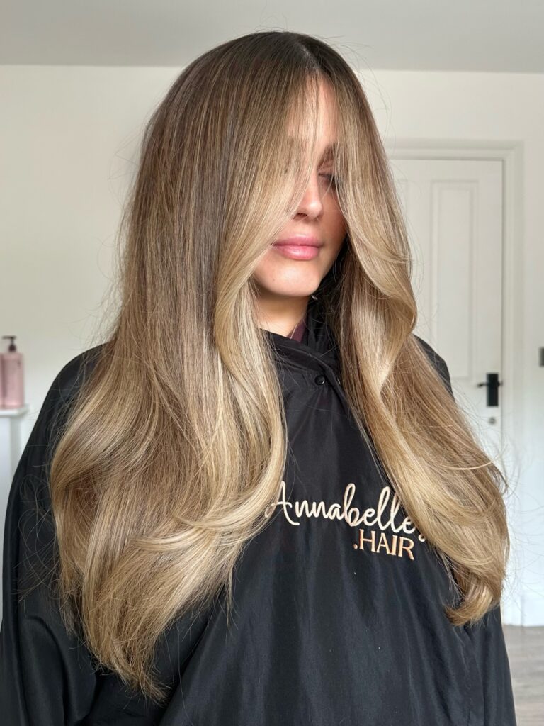 A woman with long, straight blonde balayage hair featuring wispy face-framing layers.