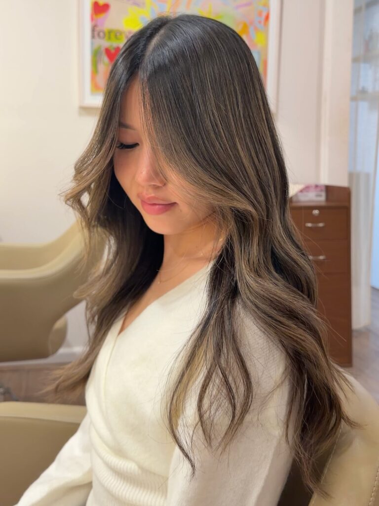 A woman with long wavy hair featuring dark roots that fade into caramel balayage highlights.