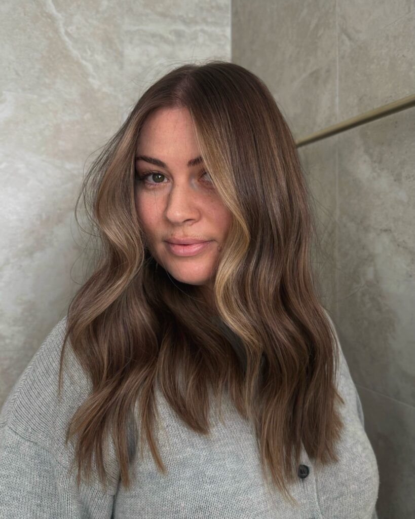 A woman with long wavy brown hair featuring soft sunkissed highlights around the face.
