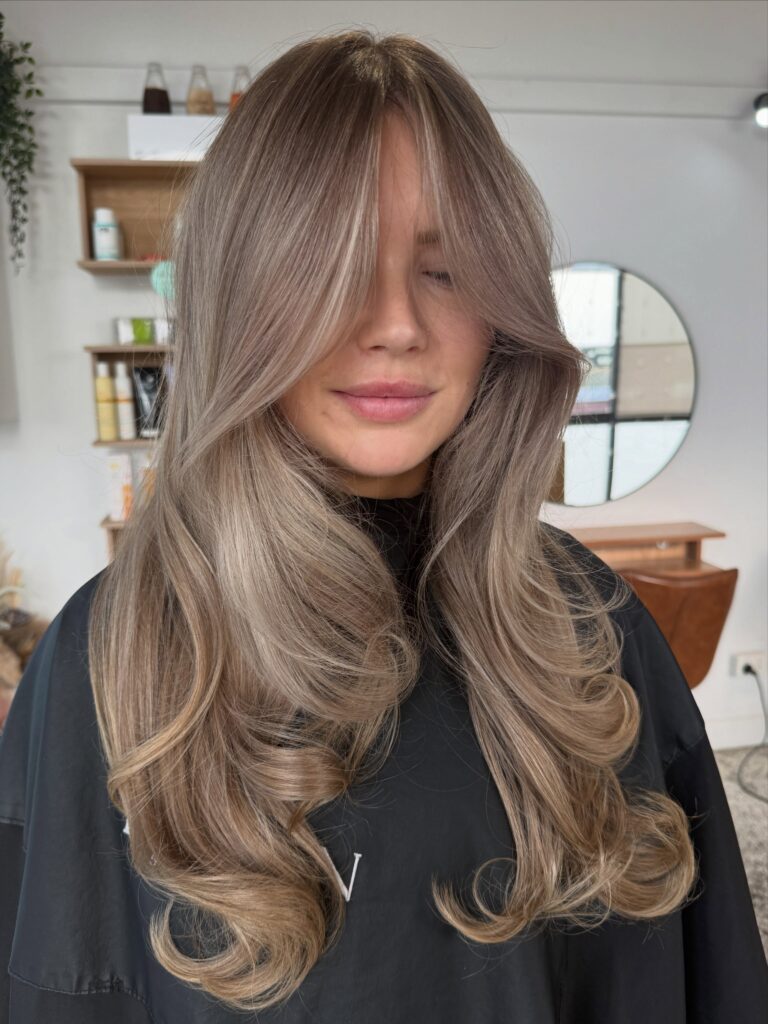 A woman with long ash blonde layered hair featuring soft curtain bangs and curled under ends.