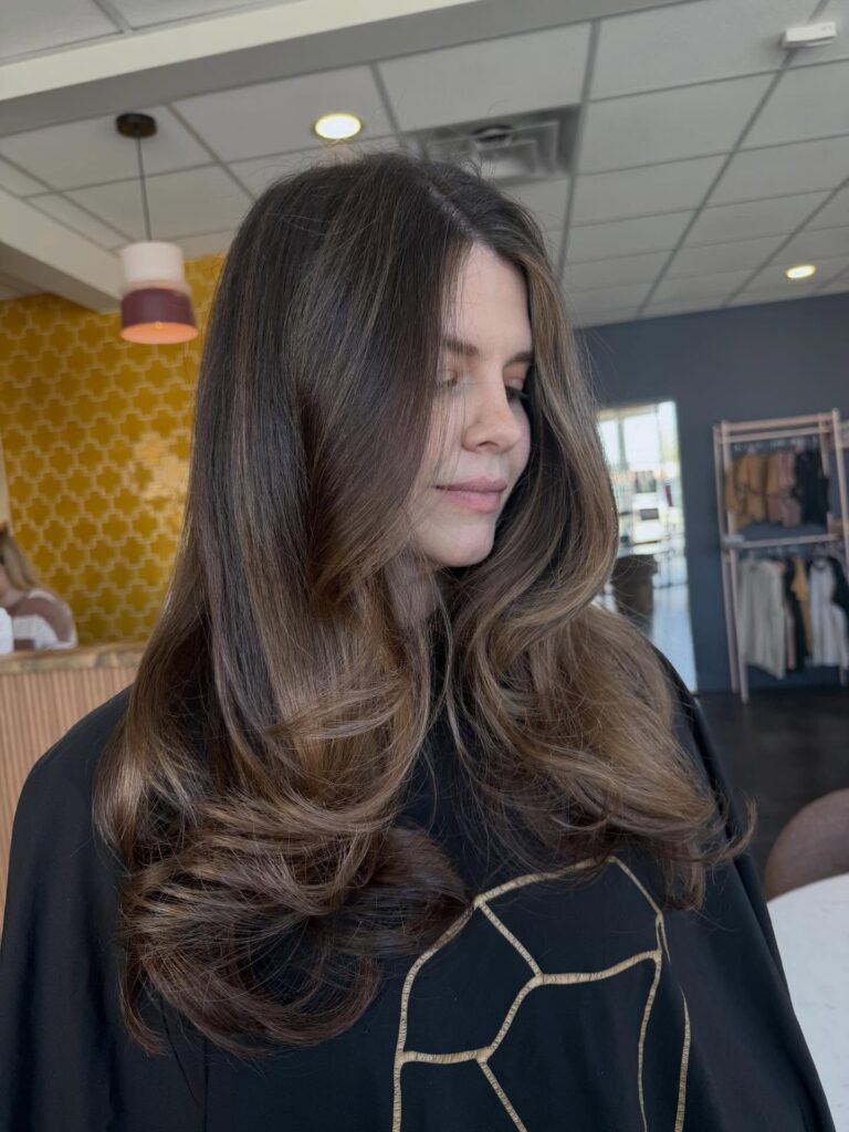 A woman with long, glossy chocolate brown hair featuring soft flipped-out layers and subtle highlights.