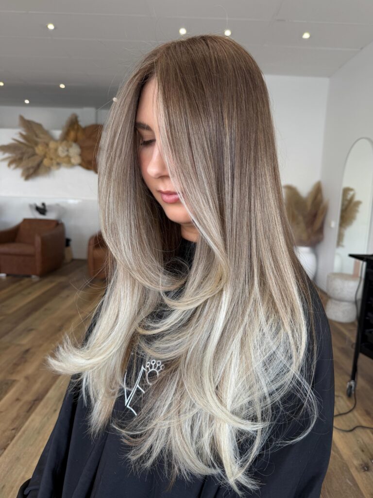A woman with long straight hair featuring a dark rooted top that fades into icy blonde ends.