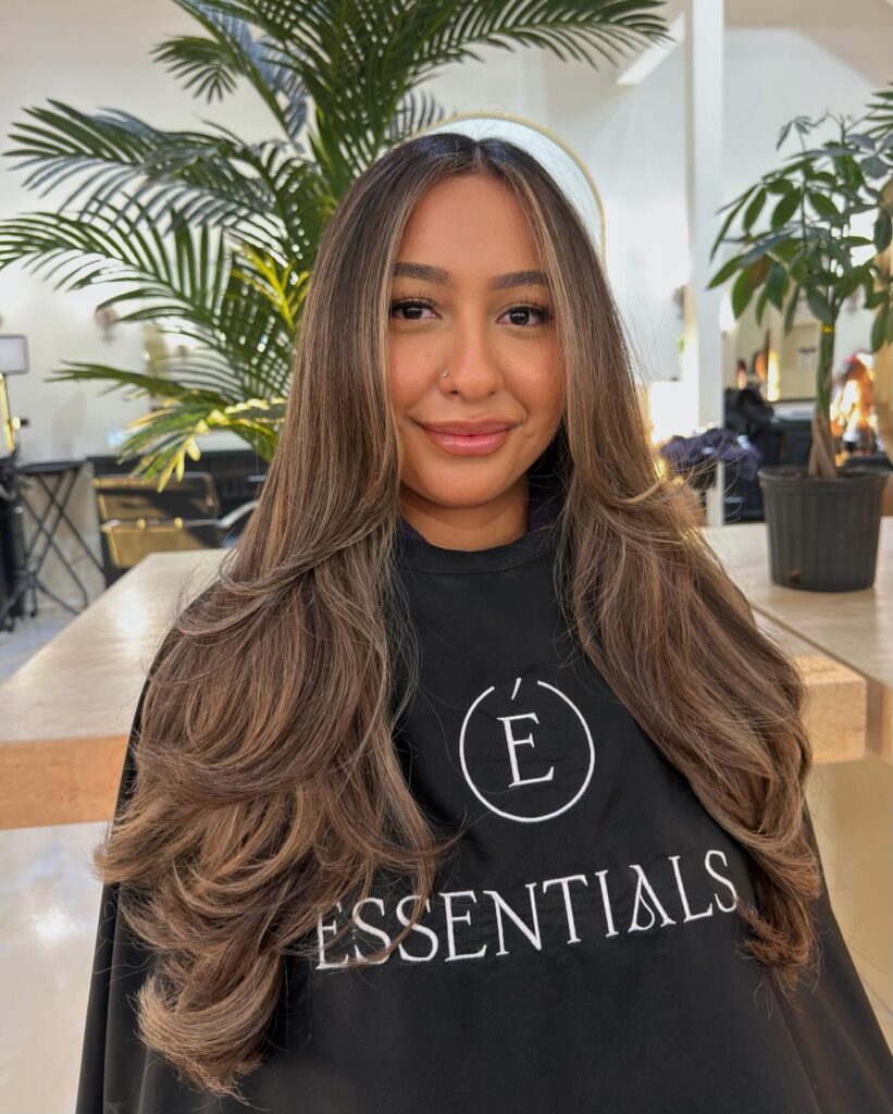A woman with long, layered brunette hair featuring caramel balayage highlights and soft curled ends.