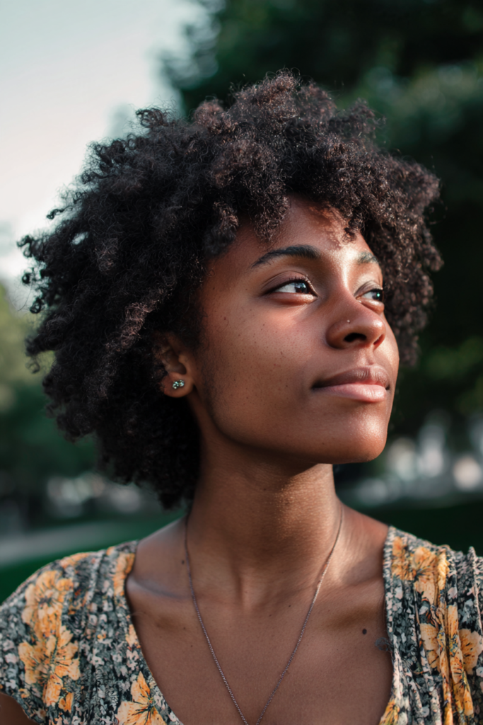 A woman with a chin length curly bob hairstyle smiles softly while looking to the side outdoors.
