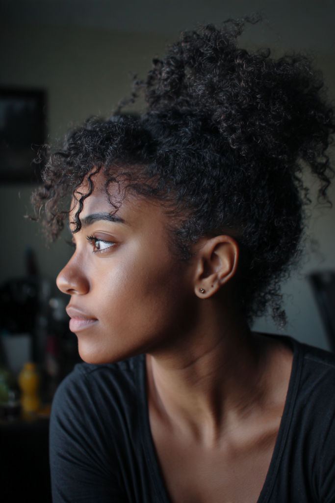 A woman with a curly bun and loose curtain bangs looks off to the side while wearing a dark top.