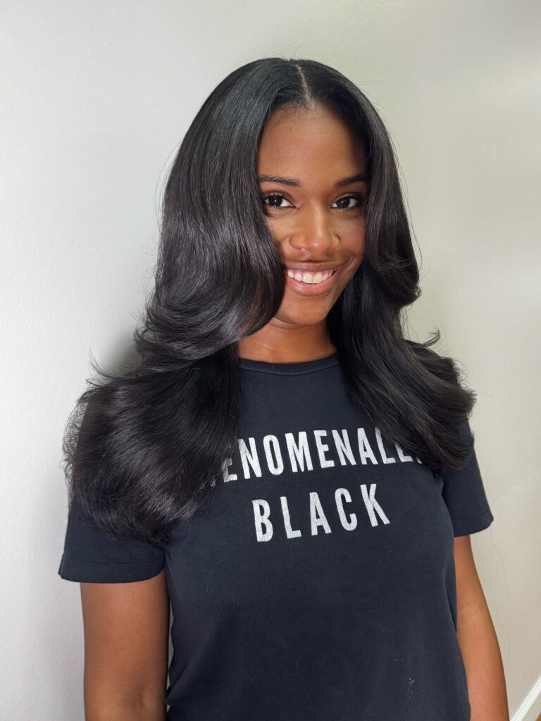 A woman with a long silky middle part blowout hairstyle smiles while wearing a black graphic t-shirt.