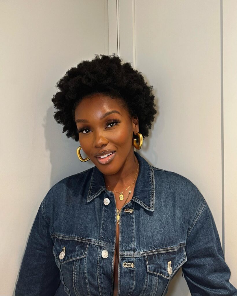 A woman with a short natural twist out hairstyle featuring tight coily texture and a rounded silhouette smiles while wearing a denim jacket and gold hoop earrings.