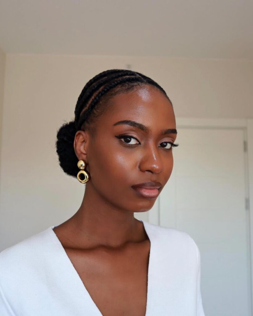 A woman with sleek cornrow braids leading into a low bun wears gold statement earrings and a white top.