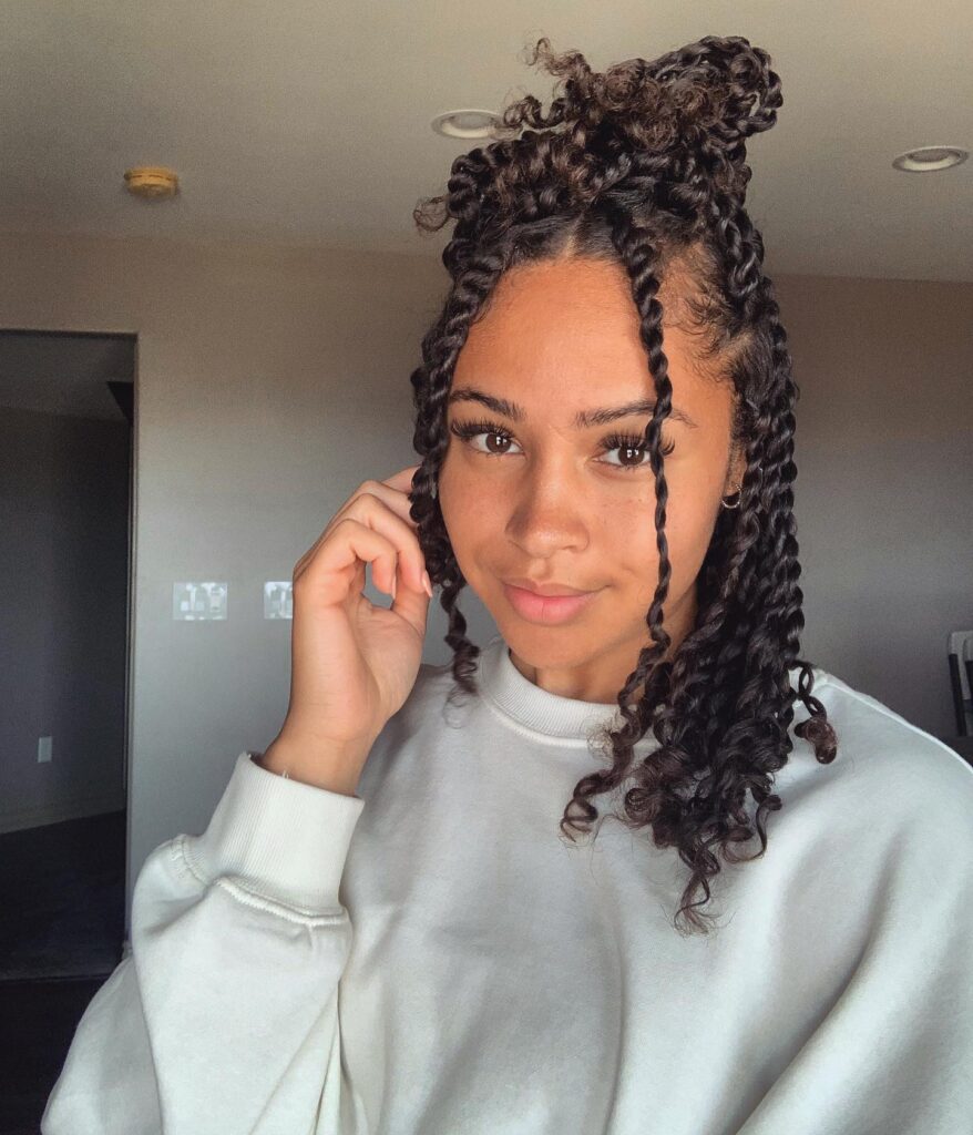A woman with long dark twisted hair styled into half up space buns smiles while wearing a white sweatshirt indoors.