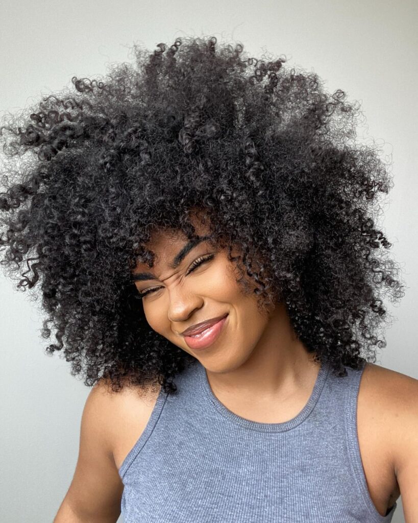 A woman with a voluminous curly shag hairstyle laughs while wearing a gray tank top against a light background.