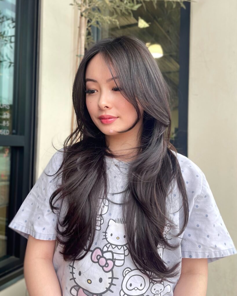 A woman with long dark brown hair featuring a deep side part, curtain bangs, and soft curled layers.