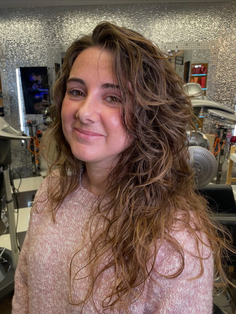 A woman with long curly light brown hair featuring caramel highlights and heavy layers.