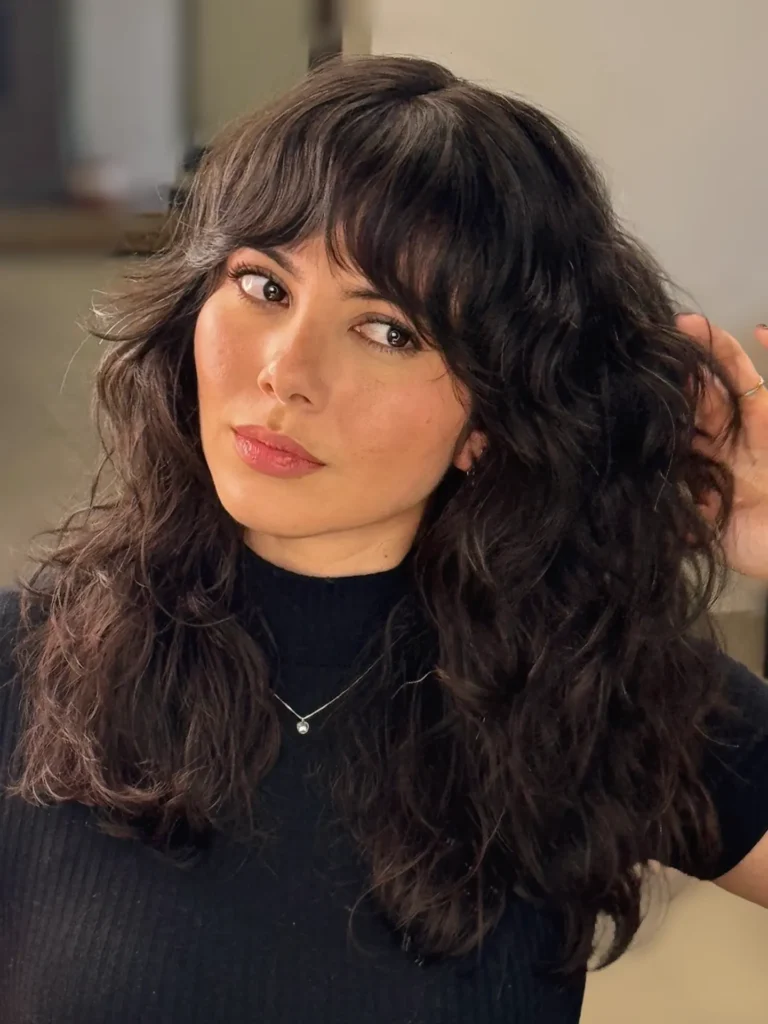 A woman with dark brown voluminous curly layered hair and wispy curtain bangs.