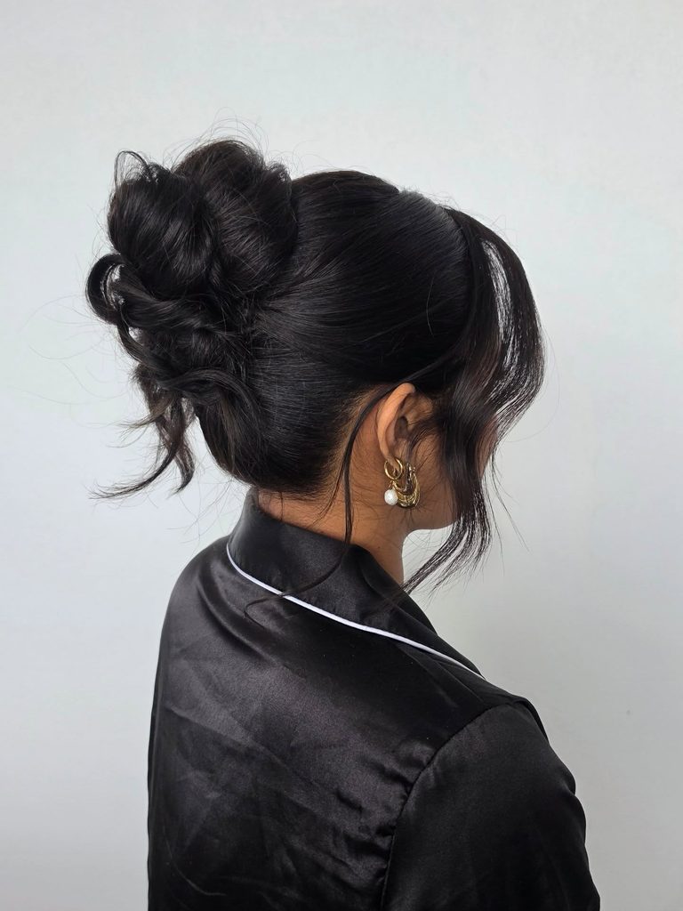 Side view of a woman with glossy black hair styled in a smooth high crown with a loose twisted bun, soft wavy bangs and loose strands, wearing gold pearl hoop earrings and a black satin top.