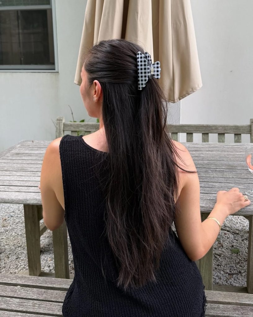 Back view of a woman with long straight dark brown hair in a half up style secured with a black and white gingham claw clip.