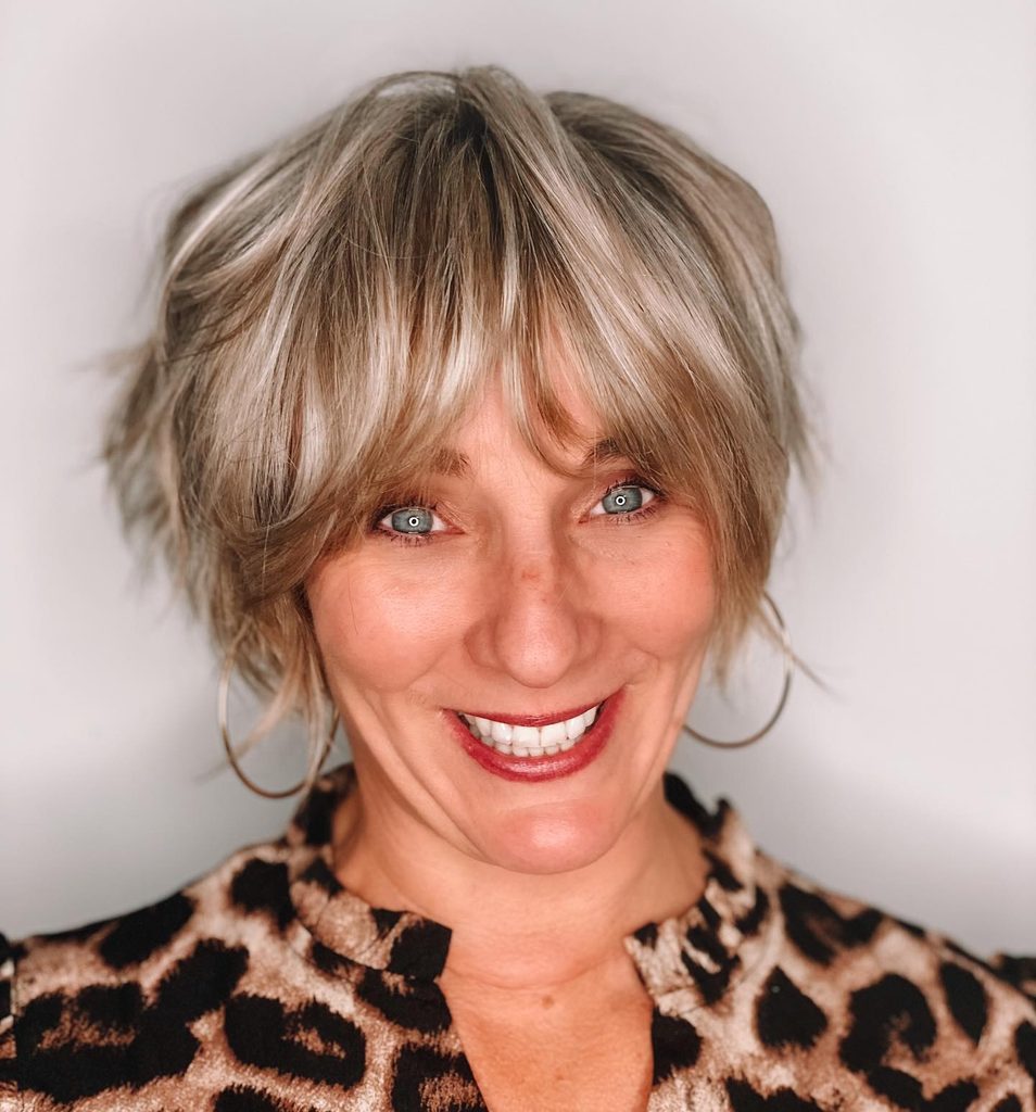Smiling woman with a short shaggy pixie bob in ash blonde and silver tones, styled with full wispy bangs and choppy, textured layers.