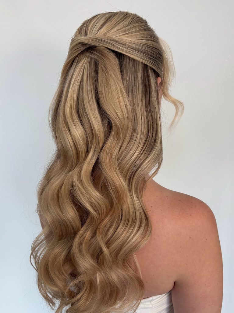 Back view of a woman with long honey blonde wavy hair styled in a half up criss cross twist with a curled strand left out at the side.