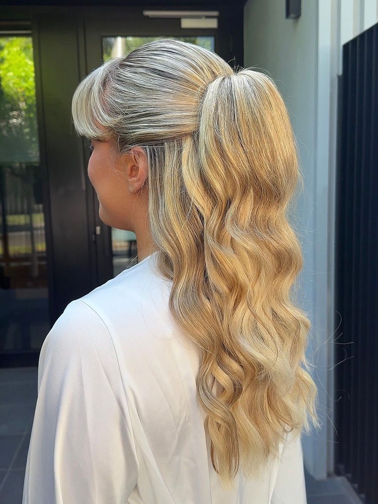 Side view of a woman with long golden blonde wavy hair and short blunt bangs styled in a half up pony at the crown.