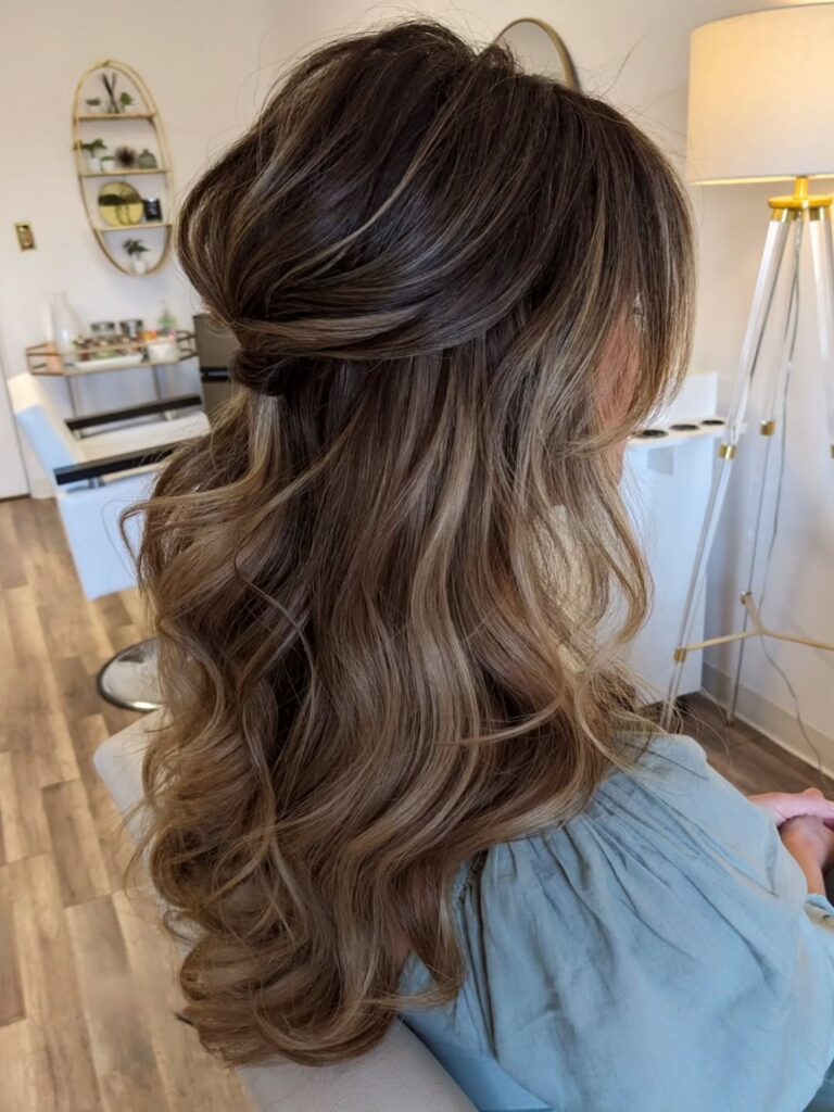 Side view of a woman with long brunette hair and caramel balayage styled in a teased half up crown with loose bouncy waves and soft face framing pieces.