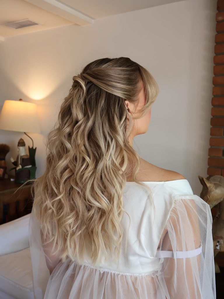 Side back view of a woman with long blonde beachy curls and side swept bangs styled in a loose half up twist at the crown.