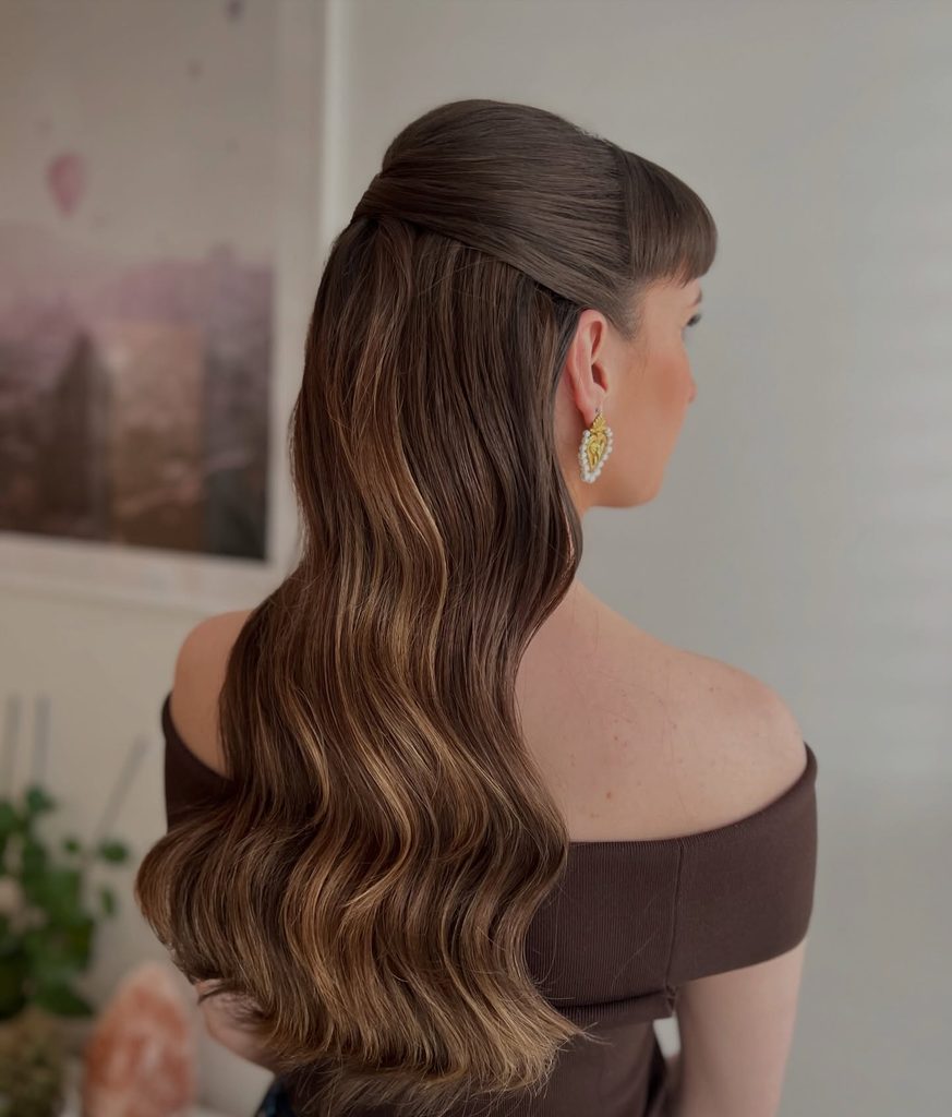 Side back view of a woman with long chocolate brown hair and short bangs styled in a sleek half up with soft caramel highlighted waves.