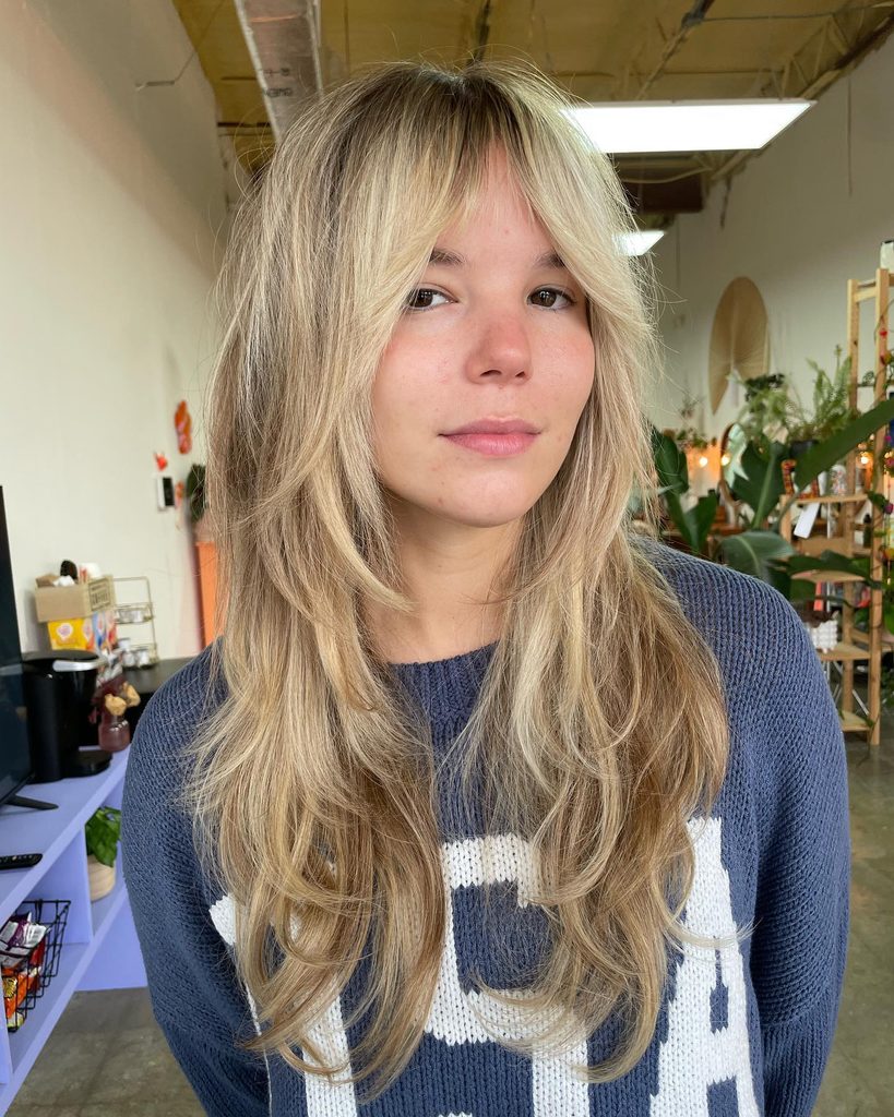 Woman with long sandy blonde shag hair, wispy curtain bangs, and soft feathered layers with a brown root smudge, wearing a blue knit sweater.
