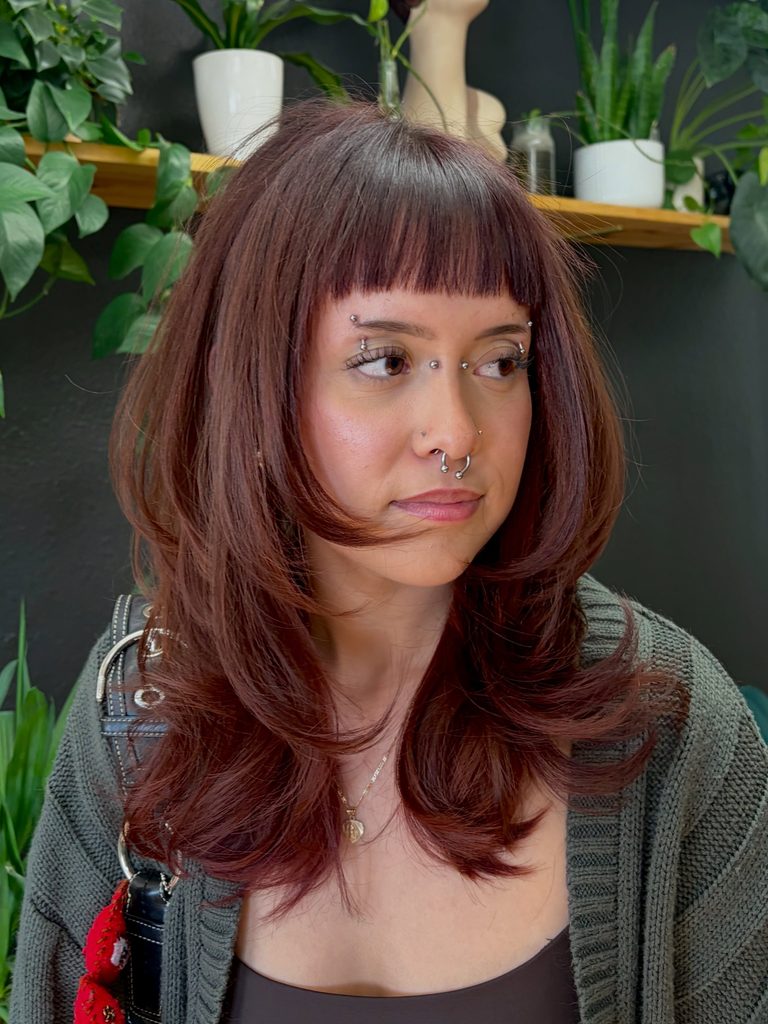 Woman with medium length cherry cola red shag hair, short blunt bangs, and sleek feathered layers, looking to the side in a gray knit cardigan with a gold pendant necklace.
