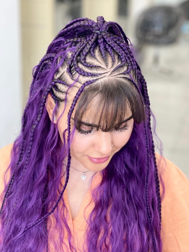 Woman with long vivid purple crimped waves, flower-patterned cornrow braids on top, thin side braids and full dark bangs, wearing an orange top and a heart pendant necklace.
