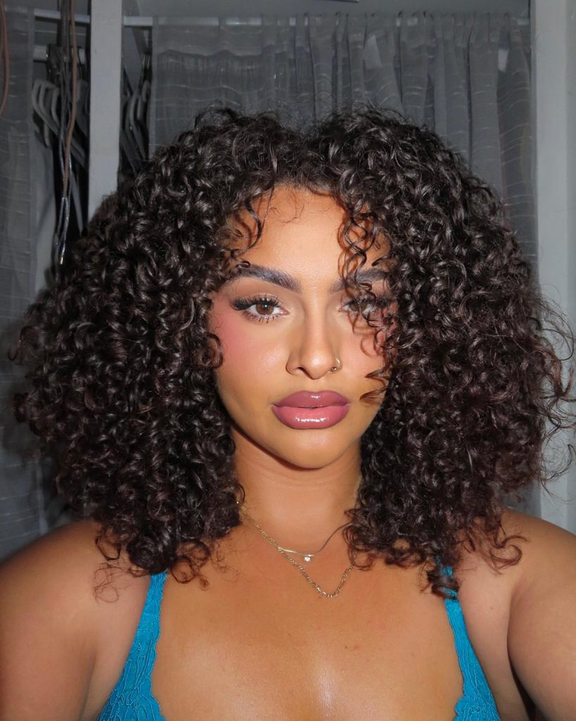 A woman with shoulder-length dark brown defined curls, rounded layers and side swept curly bangs wears a turquoise lace top and a gold necklace.