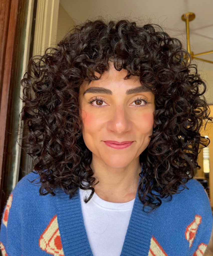 A smiling woman with collarbone-length dark brown corkscrew curls, rounded layers and textured curly fringe wears a blue patterned cardigan.