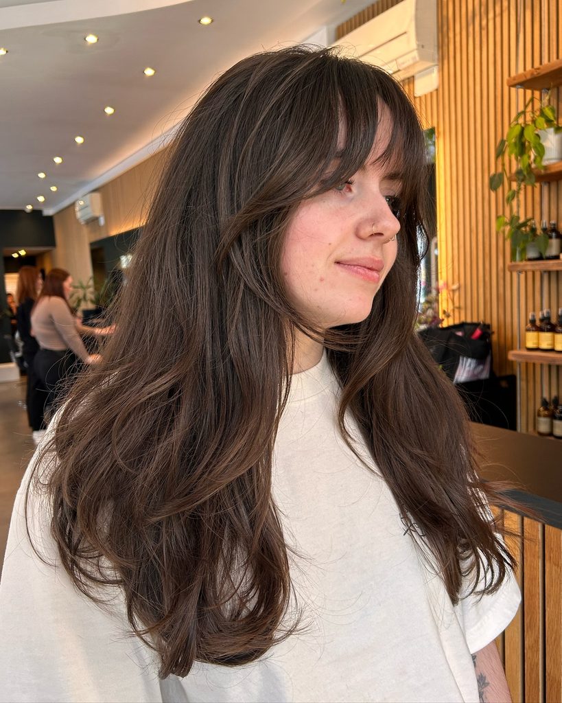 Woman with long mocha brown layered hair, soft center parted curtain bangs and loose bouncy ends with a glossy finish.