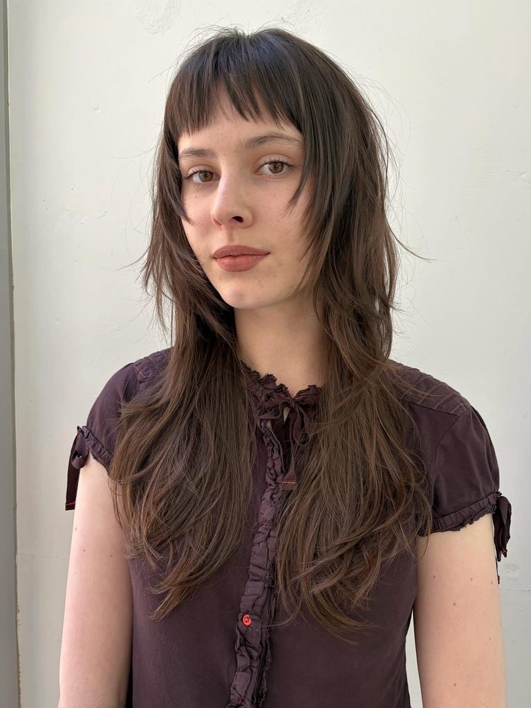 Woman with long chestnut brown shaggy layered hair, short wispy bangs and choppy face framing pieces in a straight, textured style.