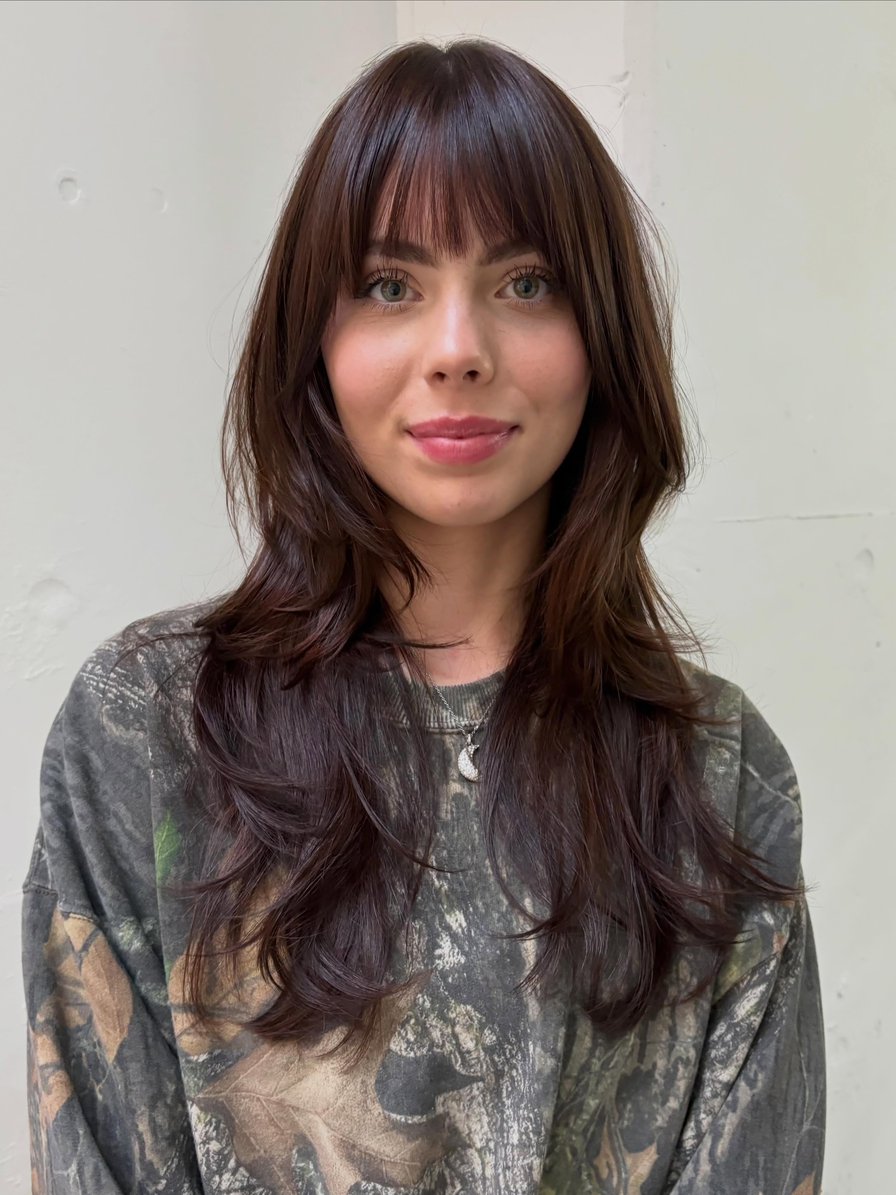 Woman with long dark brown layered hair with a subtle burgundy tint, full center parted bangs and soft feathered ends with a glossy finish.