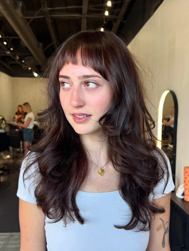 Woman with long, voluminous dark cherry brown curled layers and short micro bangs wearing a light blue top and a gold heart necklace.