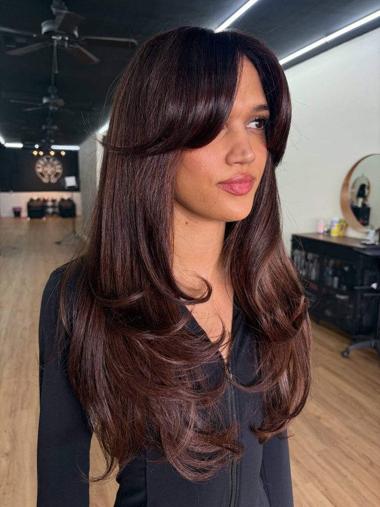 Woman with long glossy mahogany brown layered hair styled with side-swept curtain bangs and soft flicked ends in a smooth, shiny finish.