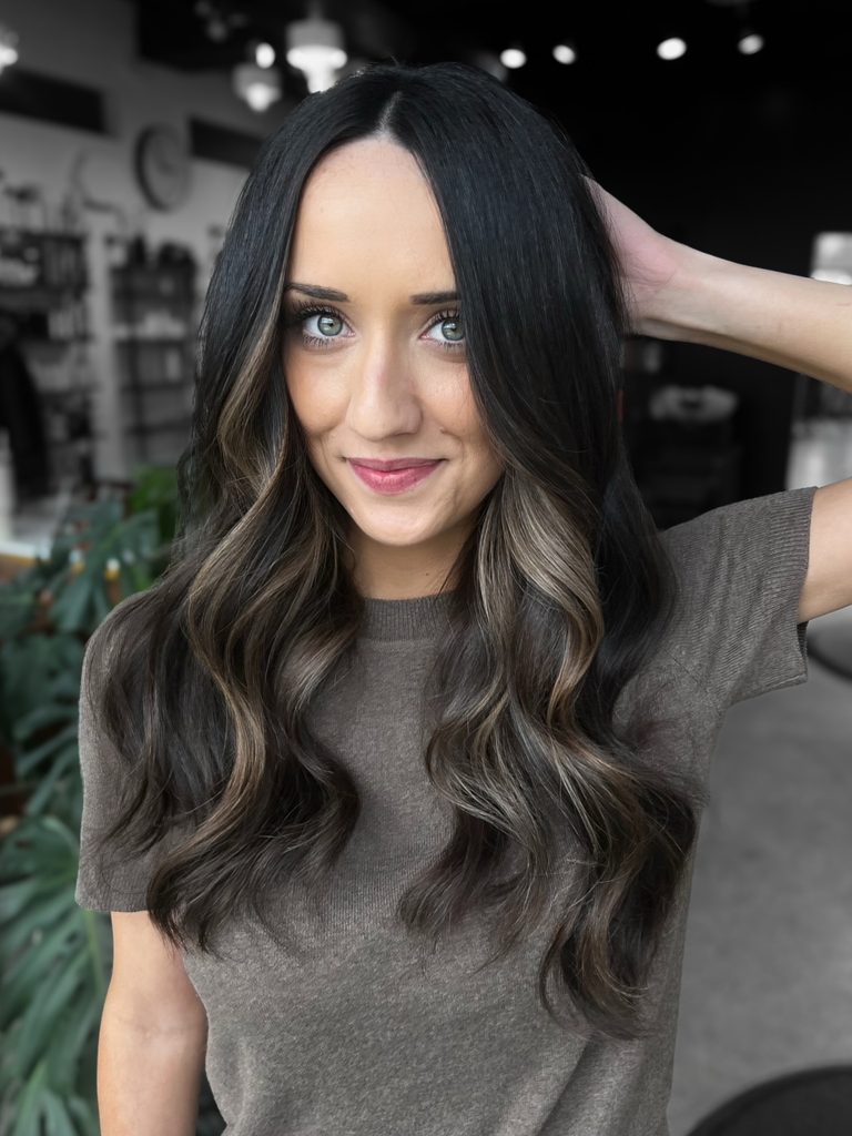 Woman with long dark brown hair parted in the center and styled in loose glossy waves with soft honey blonde highlights framing her face and blending through the ends.