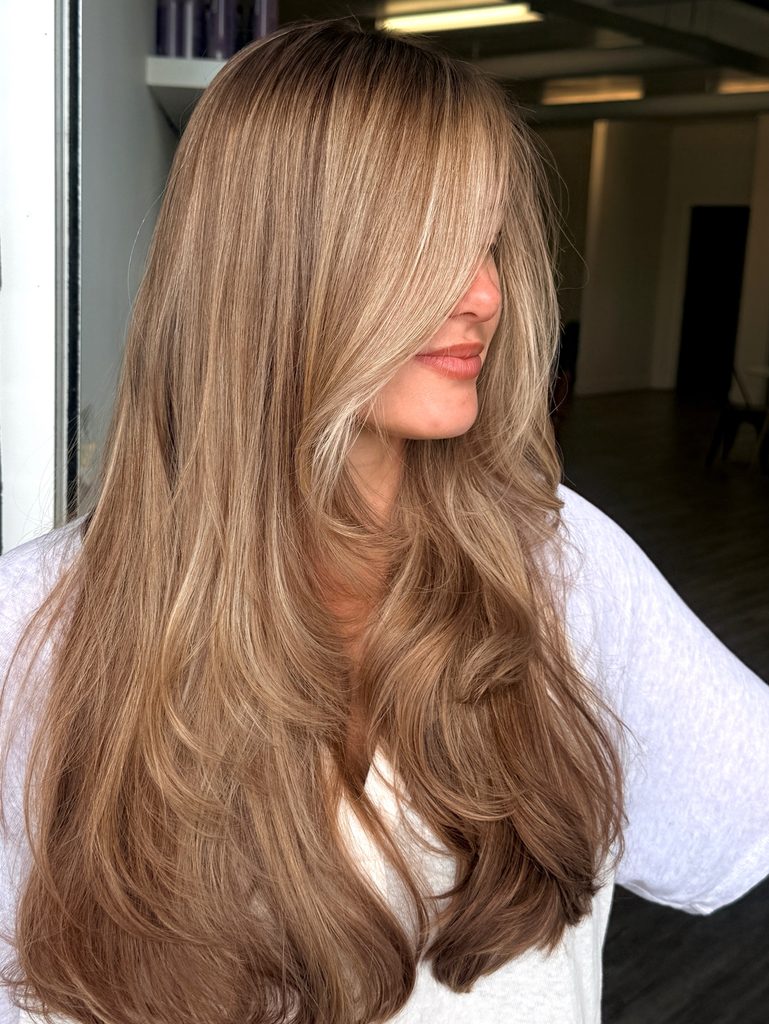 Woman with long layered caramel brown hair with creamy blonde face-framing highlights, long side-swept bangs and smooth inward-curved ends in a glossy blowout finish.