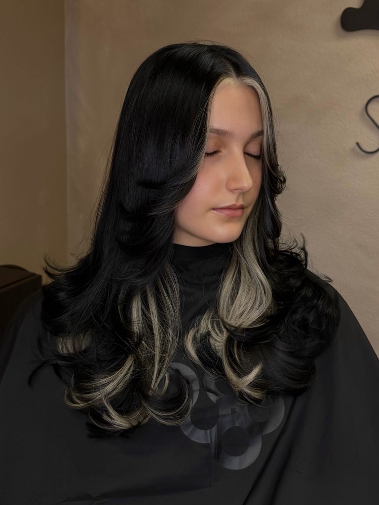 Woman with long black layered hair styled in bouncy outward curls with a center part and bright platinum blonde money piece highlights framing her face and woven through the ends.