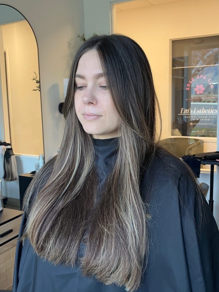 Woman with long straight dark brown hair parted in the center with face-framing layers and soft caramel blonde highlights throughout the lengths and ends.