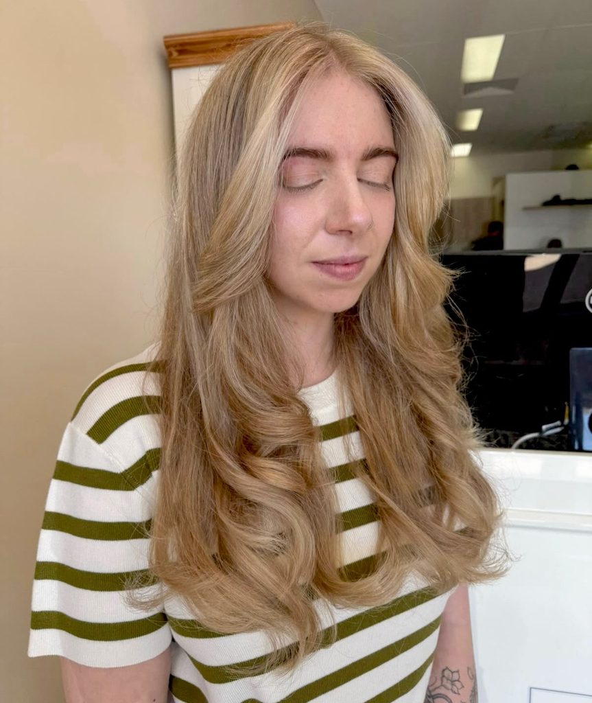 Woman with long golden honey blonde hair parted in the center and styled in soft layers with loose flowing waves and a warm blended color.