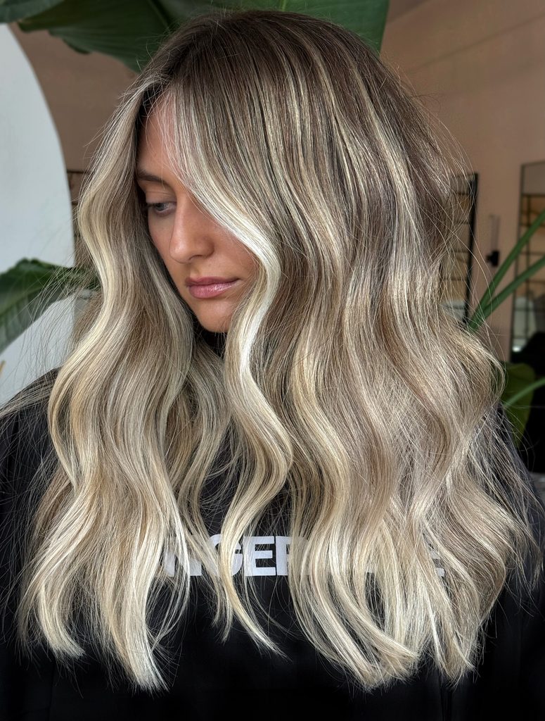 Woman with long wavy bronde hair parted in the center and styled in loose beachy waves with a shadowy brown root and bright creamy blonde highlights framing her face and flowing through the ends.