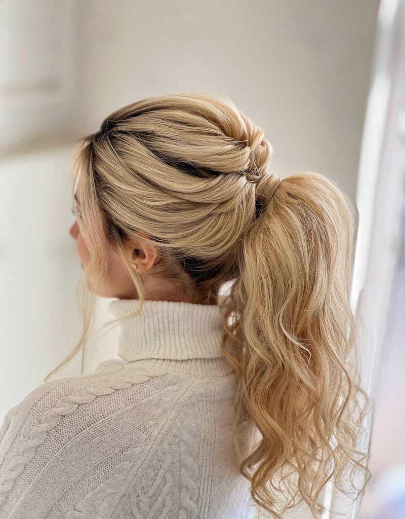 Back view of a woman with a high ponytail of long, wavy honey blonde hair, a twisted voluminous crown, and wispy bangs near her ear.