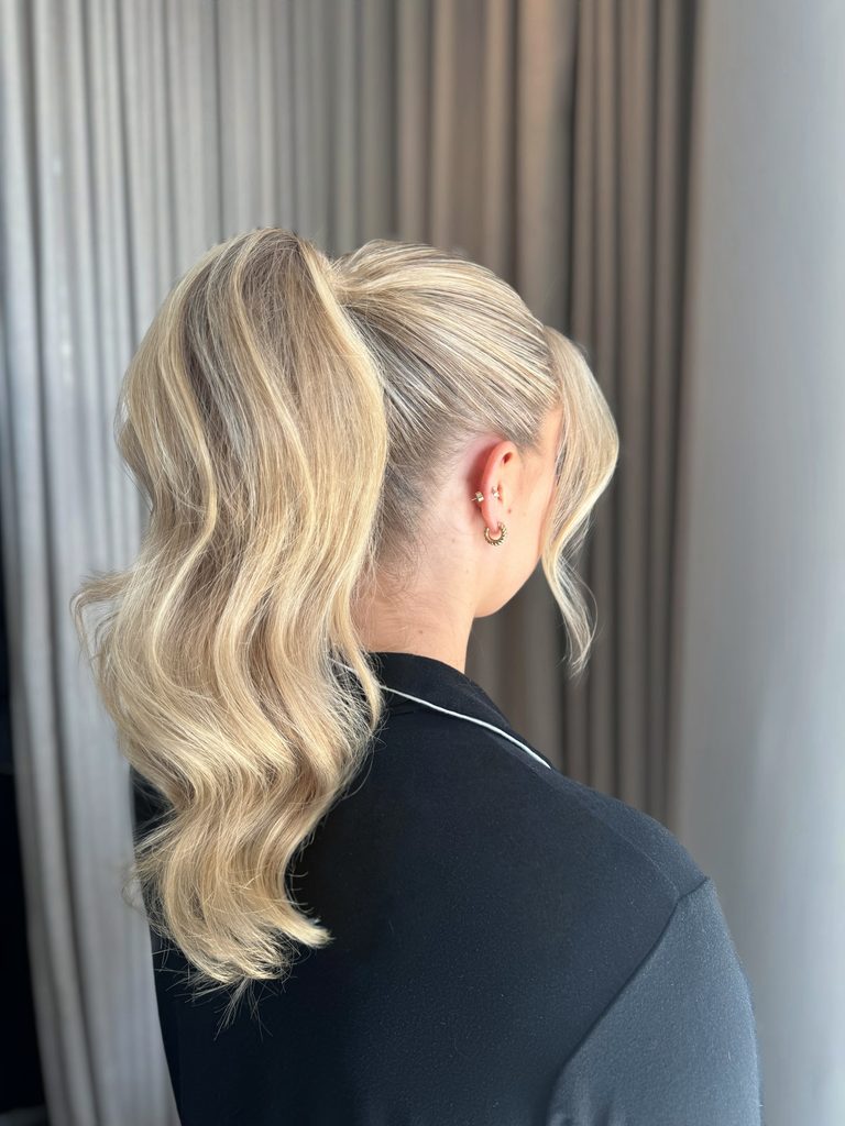 Back view of a woman with a high ponytail of long, wavy buttery blonde hair with highlights and a soft curled side bang near her ear.