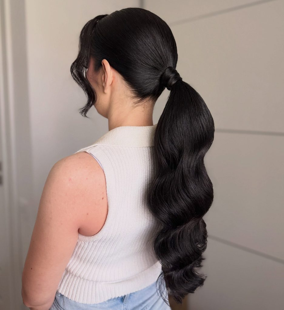 Back view of a woman with a sleek low ponytail of long, glossy jet black hair in Hollywood style waves with a curled side bang near her ear.