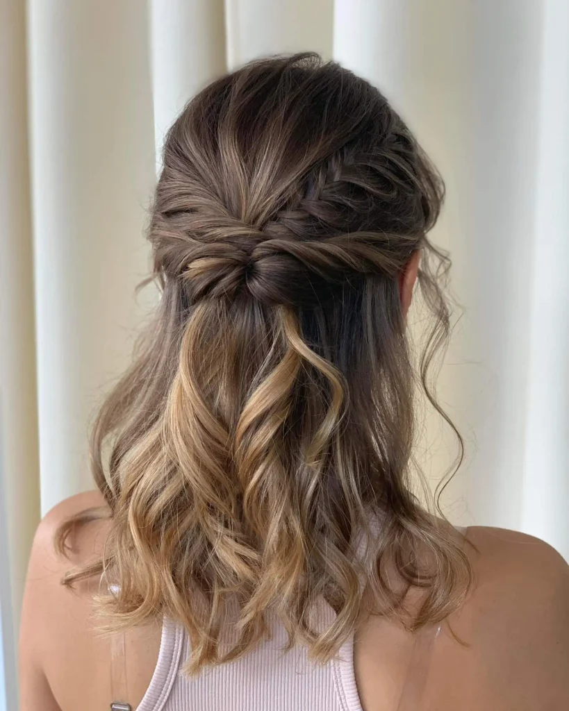 A woman with shoulder-length wavy brunette and blonde hair has a beautiful boho half-up braid and twist seen from behind.