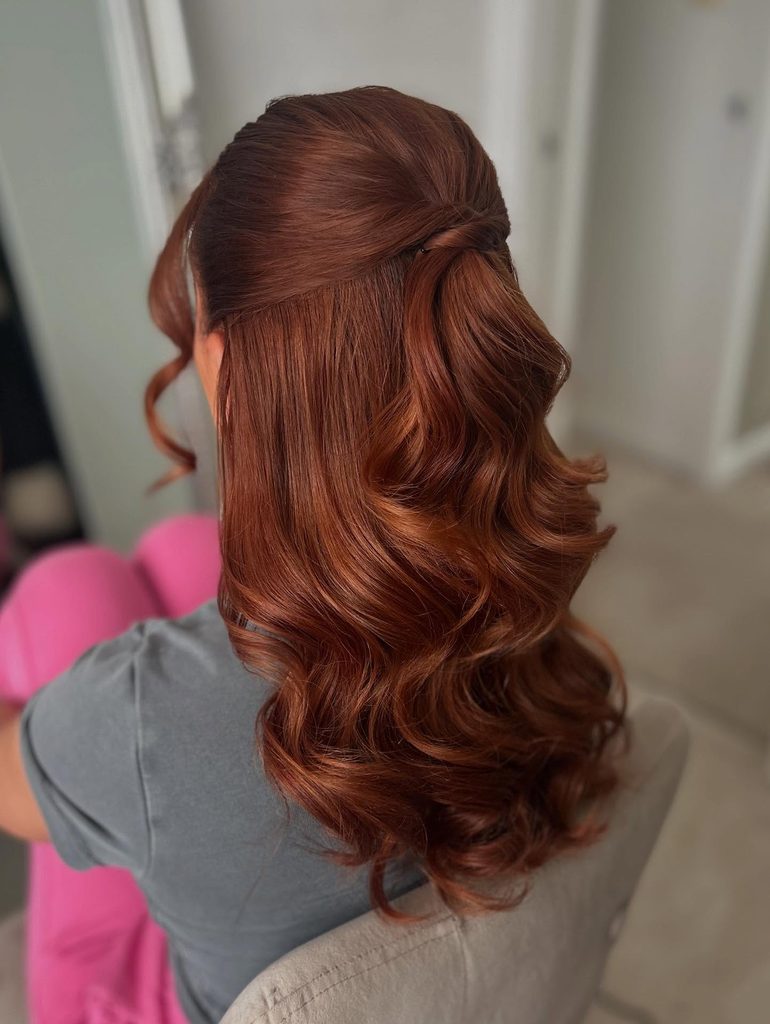 Back view of a woman with long copper red hair styled in a smooth half up style with big glossy vintage curls and a curled face framing strand.